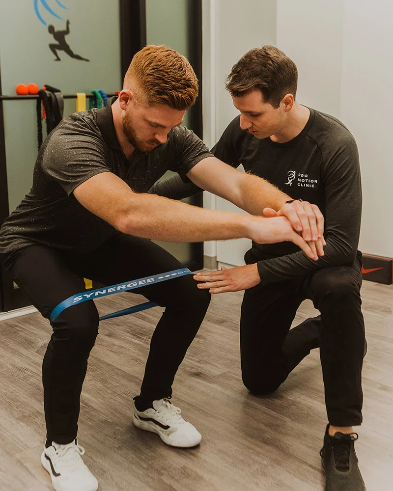 Kinesiologist supervising resistance band squat for lower body strength at Pro Motion Clinic