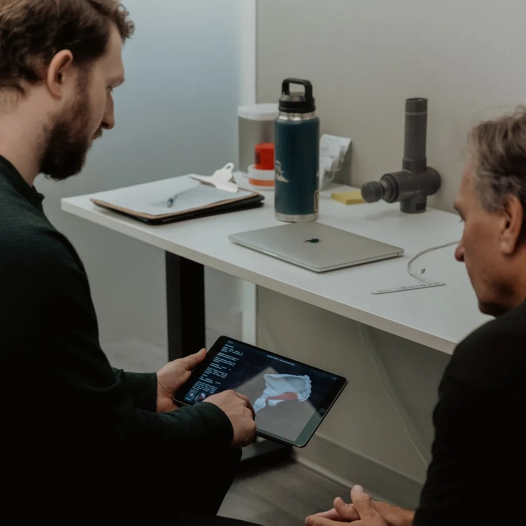 Chiropractor reviewing 3D joint anatomy on tablet with veteran patient during medical assessment consultation in Kelowna