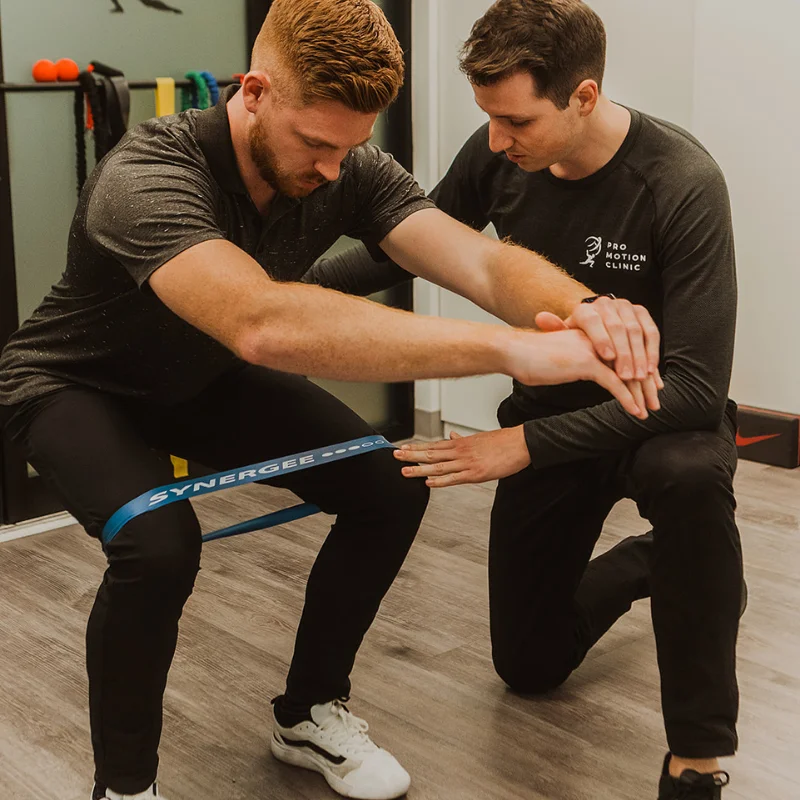Pro Motion Clinic practitioner guiding patient through resistance band squat for chronic pain rehabilitation in Kelowna